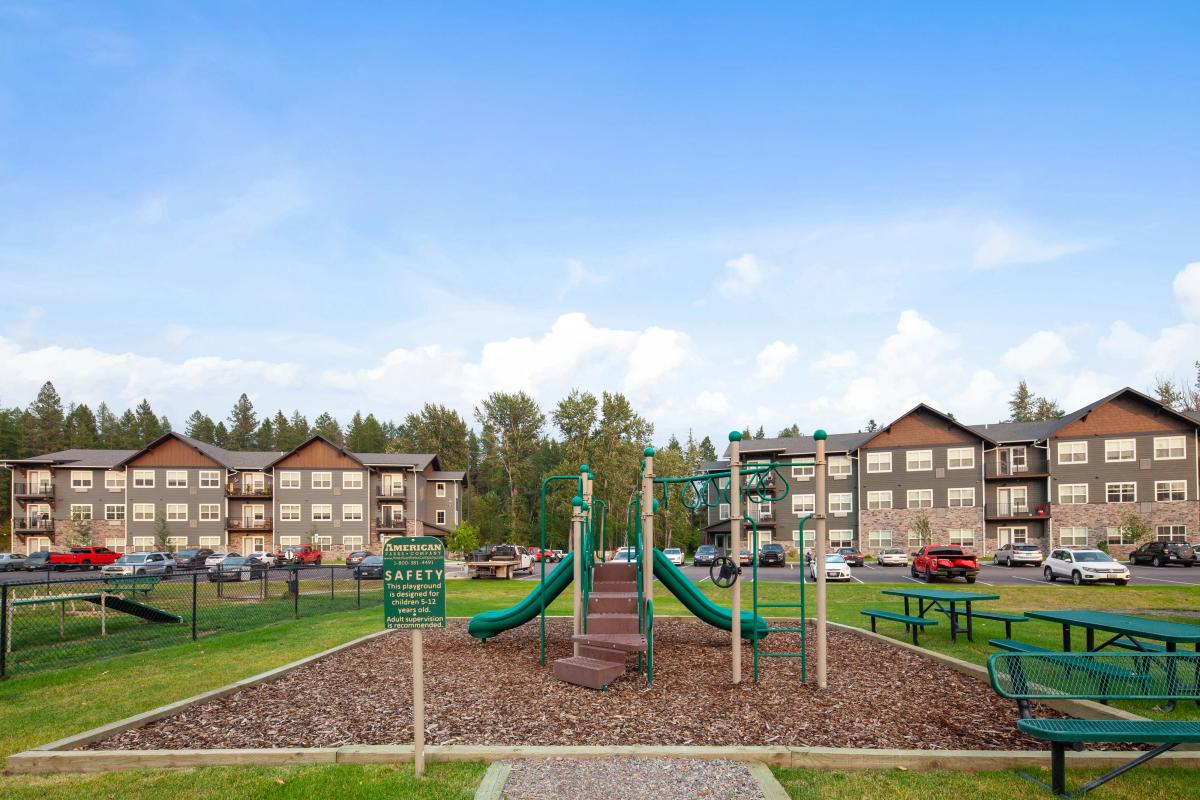 Apartment complex with a children's playground in front, surrounded by lush greenery and blue skies.